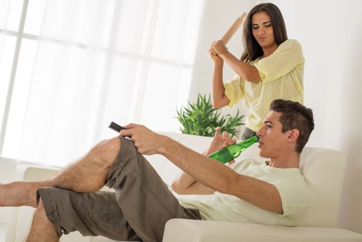 Young man sitting on sofa and watching tv. His wife is angry and standing with rolling pin.