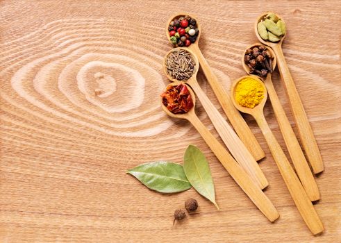 A set of several spices in wooden spoons on wooden table. In the set has a mixture of different peppers, allspice, turmeric, clove, cumin, cardamom, paprika, Bay leaf. Top view.