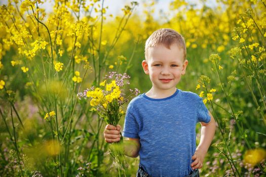Little happy boy with a bouquet of flowers is in the yellow flowering rapeseed field