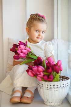cute little girl in a white dress sitting on a windowsill with a bouquet of pink tulips