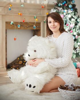 Young woman sitting on the floor in a sweater and hugs a big white teddy bear in Christmas decorations