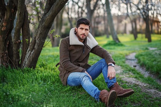 Portrait of trendy young attractive man sitting on green grass in a grove at sunset