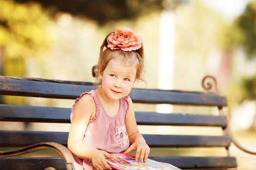 Portrait of a clever little girl reading a children's book on a park bench