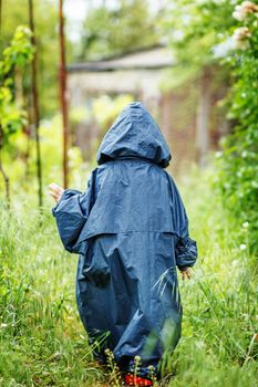 Funny little boy walks in the rain in a raincoat with a hood. Back view