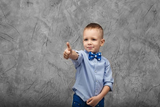 Portrait of a cute little boy in jeans, blue shirt and bow tie on a gray textural background in studio. Kid shows thumb