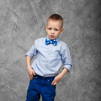 Portrait of a cute little boy in jeans, blue shirt and bow tie on a gray textural background in studio