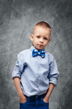 Portrait of a cute little boy in jeans, blue shirt and bow tie on a gray textural background in studio