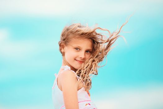 Portrait of a pretty little girl with waving in the wind long hair sitting on the beach against the blue sky