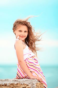 Portrait of a pretty little girl with waving in the wind long hair sitting on the beach against the blue sky