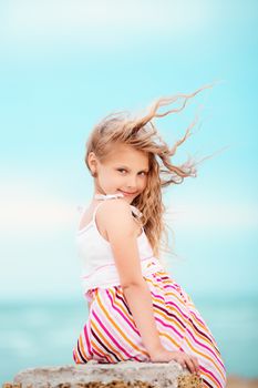 Portrait of a pretty little girl with waving in the wind long hair sitting on the beach against the blue sky