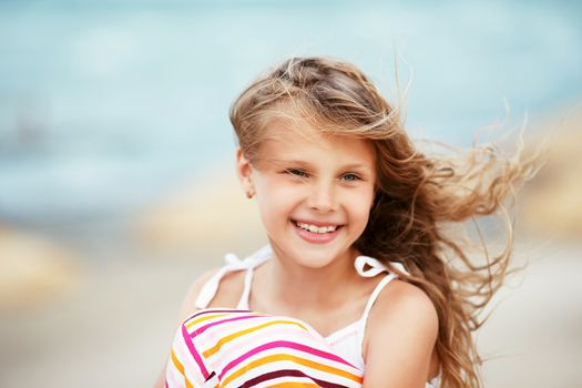 Portrait of a pretty little girl with waving in the wind long hair sitting on the beach