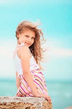 Portrait of a pretty little girl with waving in the wind long hair sitting on the beach against the blue sky