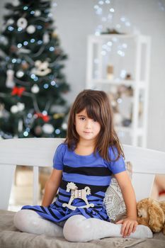 Portrait of pretty brown-eyed little girl in a blue striped dress sitting on a white bench in New Year's interior