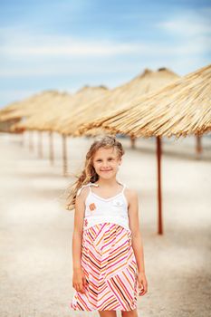 Portrait of a beautiful little girl in a sundress standing on a golden beach among tropical bamboo umbrellas