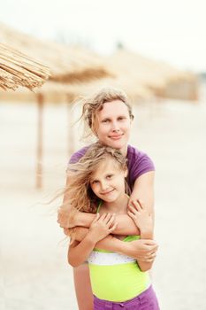 Loving mother hugging her little daughter on tropical sandy beach among the thatched umbrellas