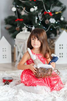 Portrait of a pretty little brunette girl holding a toy bunny in a basket in the interior with Christmas decorations