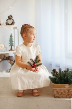 Portrait of a cute little blonde girl holding fir twig and looking out the window in the interior with Christmas decorations. Beautiful Little girl waiting for a miracle in Christmas decorations