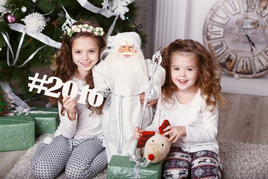 Little girls in comfortable home clothes sitting on floor in beautiful Christmas decorations. Two little sisters decorating Christmas tree with fir-cone. New year preparation. Happy girls and family.