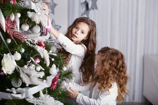 Little girls in comfortable home clothes in beautiful Christmas decorations. Two little sisters decorating Christmas tree with toys and balls. New year preparation. Happy girls and family.