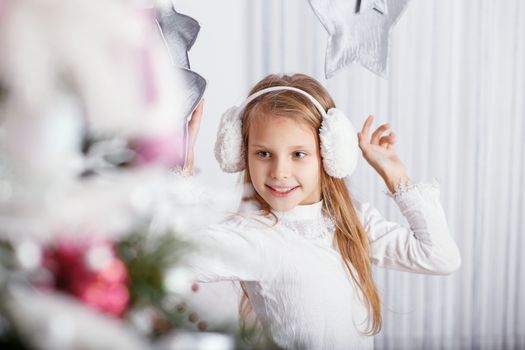 Beautiful little girl in earmuffs decorating Christmas tree with toys and balls. New year preparation.