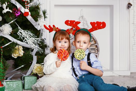 Little girl and boy in reindeer antlers eating a lollipops. Portrait of two funny little kids with a delicious candies in the hands. Christmas and New Year concept. Happy children and family In anticipation of the new year and Christmas.