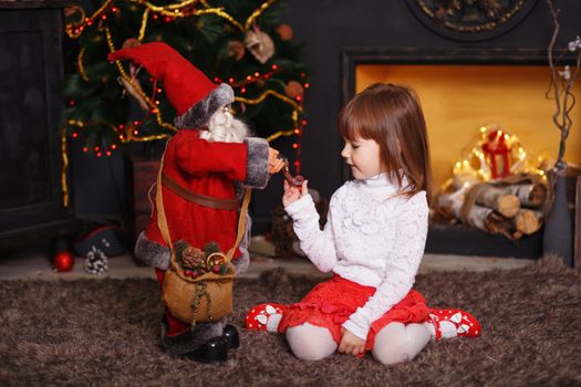 Little girl sitting on floor in beautiful Christmas decorations. Girl playing with a toy Santa Claus. New year preparation.