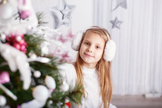 Beautiful little girl in earmuffs decorating Christmas tree with toys and balls. New year preparation.