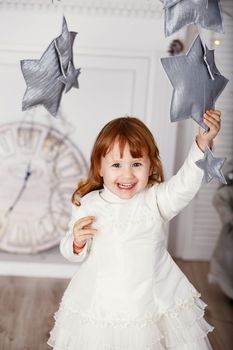 Portrait of a beautiful little girl in a white dress in the interior with Christmas decorations. Baby girl grabs and catches toy star - an element of a Christmas decor