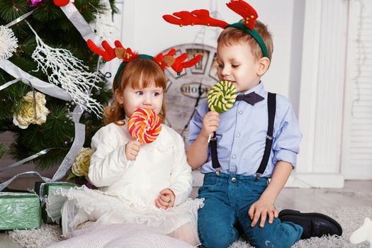 Little girl and boy in reindeer antlers eating a lollipops. Portrait of two funny little kids with a delicious candies in the hands. Christmas and New Year concept. Happy children and family In anticipation of the new year and Christmas.