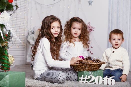 three Little kids in comfortable home clothes sitting on floor in beautiful Christmas decorations. three little siblings decorating Christmas tree with fir-cone. New year preparation. Happy girls and family.