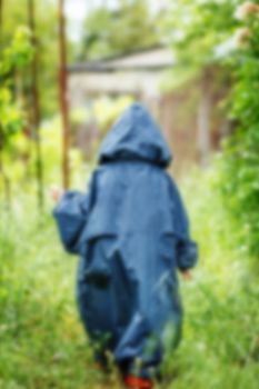 Funny little boy walks in the rain in a raincoat with a hood. Back view, blured