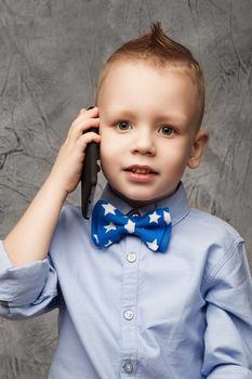 Portrait of a cute little boy in blue shirt and bow tie with mobile phone against gray textural background in studio.