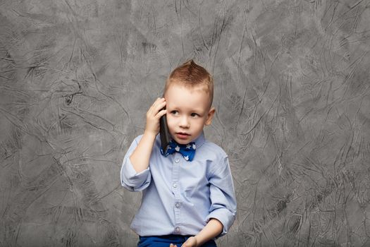 Portrait of a cute little boy in blue shirt and bow tie with mobile phone against gray textural background in studio. Business concept. Little boy businessman.