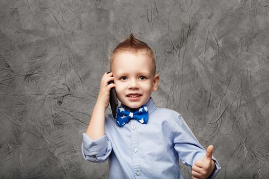 Portrait of a cute little boy in blue shirt and bow tie with mobile phone against gray textural background in studio. Kid shows thumb. Little boy American businessman.