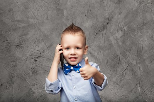 Portrait of a cute little boy in blue shirt and bow tie with mobile phone against gray textural background in studio. Kid shows thumb