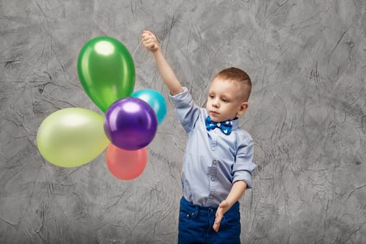 Portrait of a cute little boy in jeans, blue shirt and bow tie with multi-colored balloons in hands on a gray textural background in studio