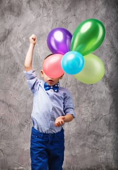 Portrait of a cute little boy in jeans, blue shirt and bow tie with multi-colored balloons in hands on a gray textural background in studio. Birthday boy