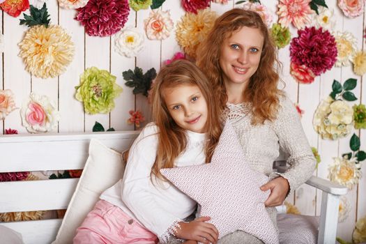 Portrait of happy smiling little girl with mother sitting on a bench against the wall decorated with flowers. Mother's Day