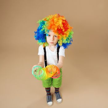 Happy clown boy with large colorful wig. Little boy in clown wig plays with a spring. Portrait of a child shot on a wide-angle lens. Birthday boy. Positive emotions. Top view portrait
