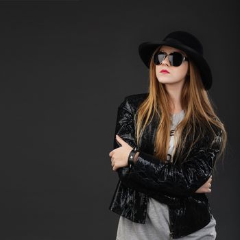 Portrait of beautiful young girl wearing black felt hat, Sunglasses and leather jacket in front of a dark gray background