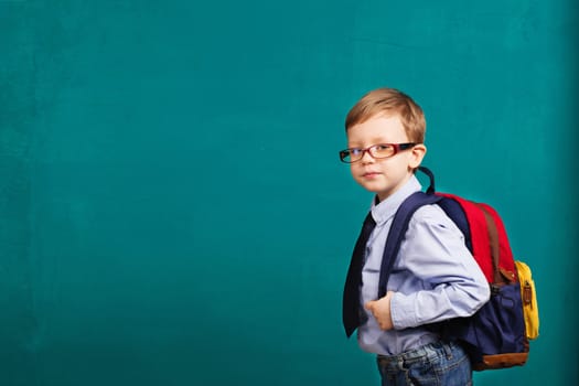 School, kid, rucksack. little Boy in eyeglasses. Cheerful smiling little kid with big backpack against chalkboard. Looking at camera. School concept. Back to School