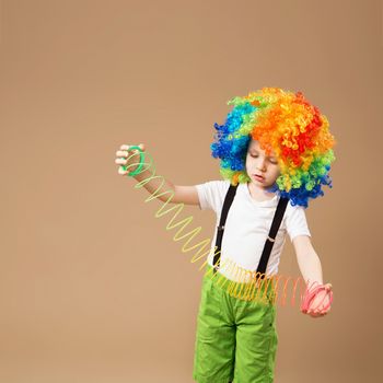 Little boy in clown wig smilling and playing with Magic Spring. Happy clown boy with large colorful wig. Birthday boy. Little clown boy with colorful hair and Slinky spring toy.