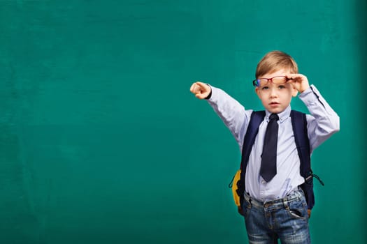 School, kid, rucksack. little Boy points a finger. Cheerful smiling little kid with big backpack against chalkboard. Looking at camera. School concept. Back to School