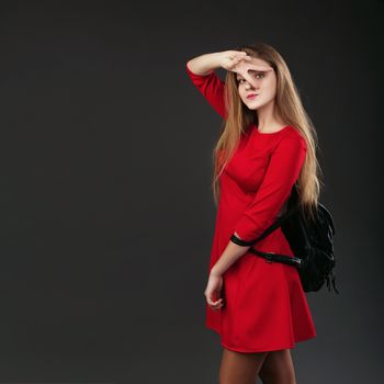 Portrait of a girl in a red dress with a black leather backpack on her shoulder. Woman with two fingers near her face