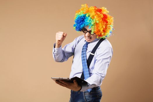 Angry office worker punches tablet. Businessman with tablet computer. Close-up Portrait of business man in clown wig using a tablet to access the internet. Business concept. Multitasking