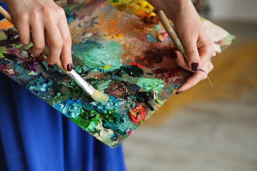 artists brushes and oil paints on wooden palette. macro artist's palette, texture mixed oil paints in different colors and saturation. palette with paintbrush and palette-knife in artist’s hands. The girl artist holding a palette with oil paints