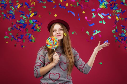 Portrait of a teen girl throws up a multi-colored tinsel and confetti. beautiful girl wearing hat holding big striped lollipop