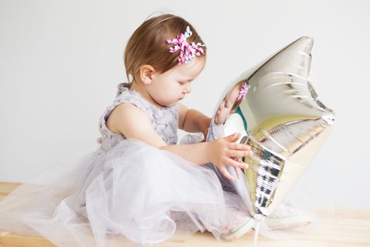 Portrait of a lovely little girl in elegant gray dress in front of a white background. Little princess. Little baby girl playing with silver star-shaped balloon.