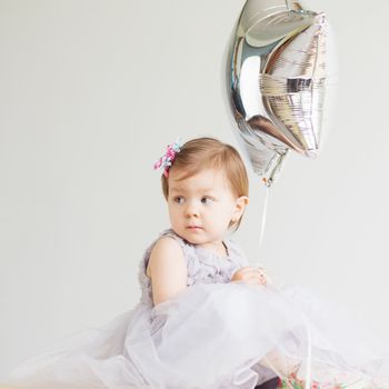 Portrait of a lovely little girl wearing elegant gray dress in front of a white background. Little princess. Little baby girl holding silver star-shaped balloon.