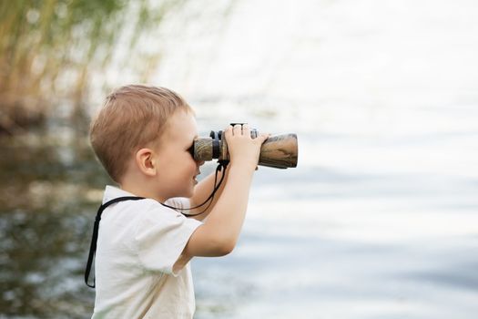 Portrait of little boy looking through binoculars on river bank. Cute kid with binoculars sitting on the pier on river